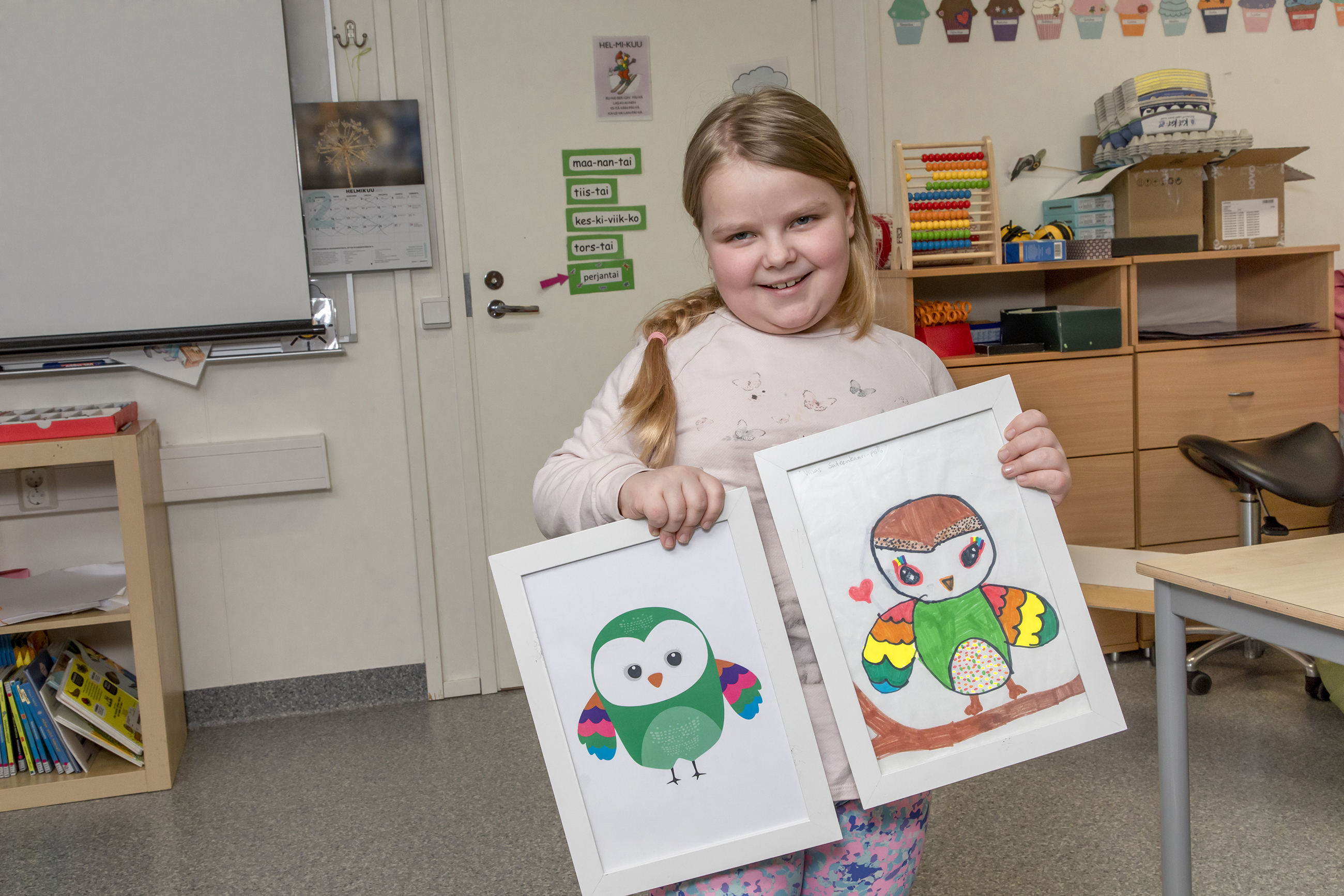 Hymyilevä Nella Lehto seisoo luokkahuoneessa ja pitää käsissään Paavali Pöllöä esittävää piirustustaan ja m...