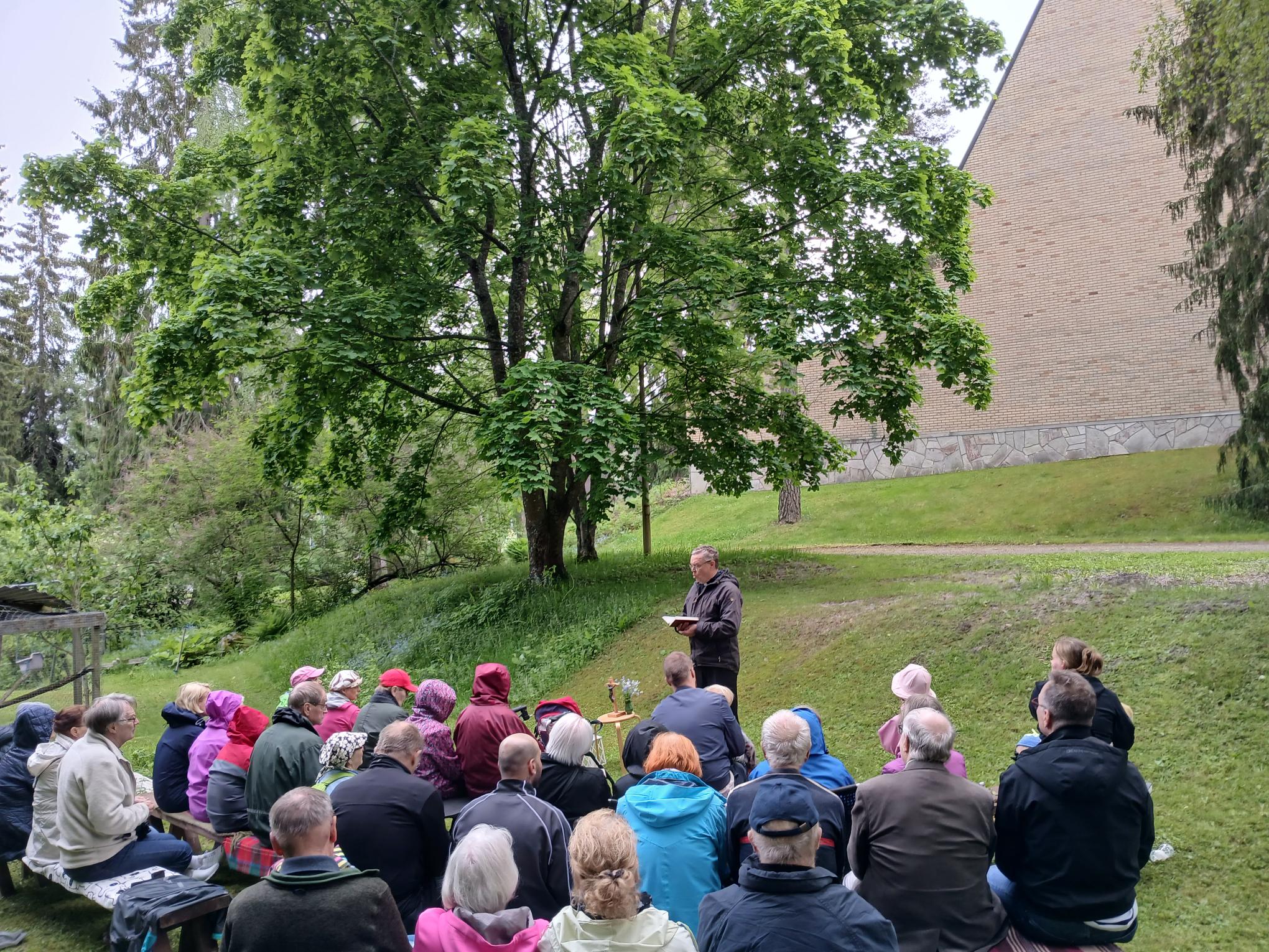 ihmisiä penkeillä, Pekka Särkiö puhumassa, taustalla vihreää luontoa ja Joutjärven kirkko.