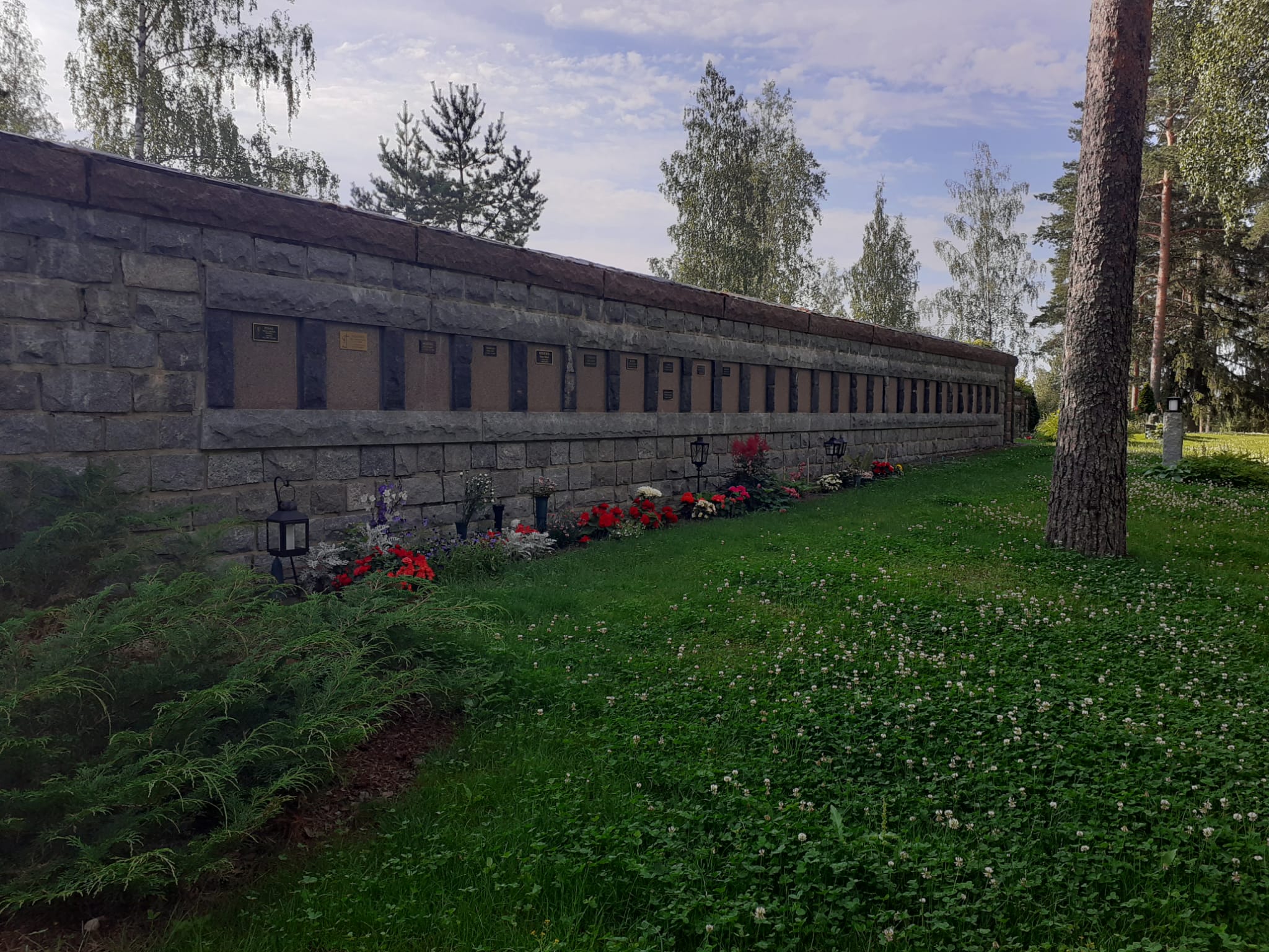 Korkea, kivinen muuri, johon kiinnitetty vainajien nimilaattoja. Muurin juurella omaisten tuomia kukkia.