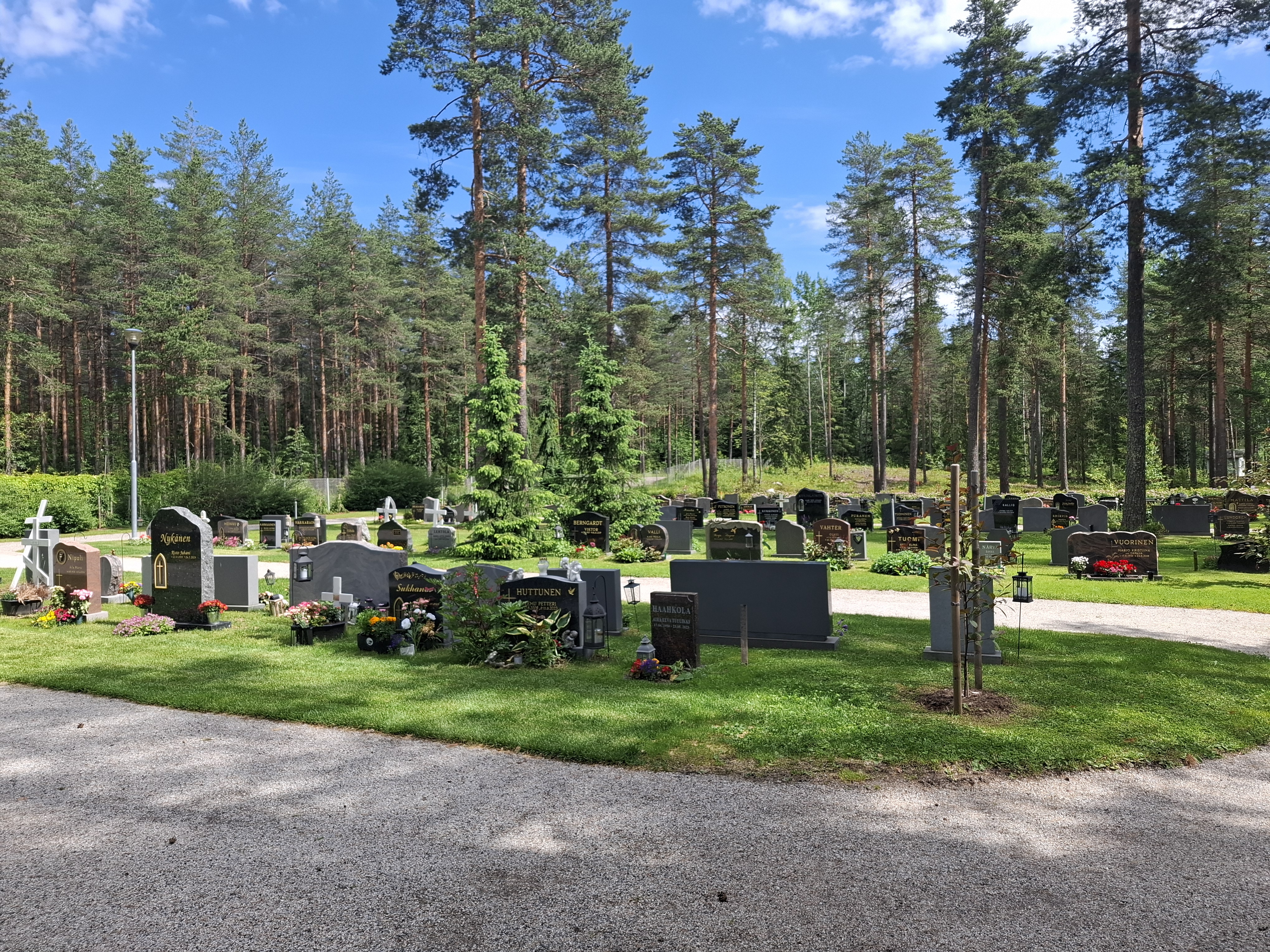 Hautamuistomerkkejä nurmikolla, leveät hiekkakäytävät välissä, taustalla puita.