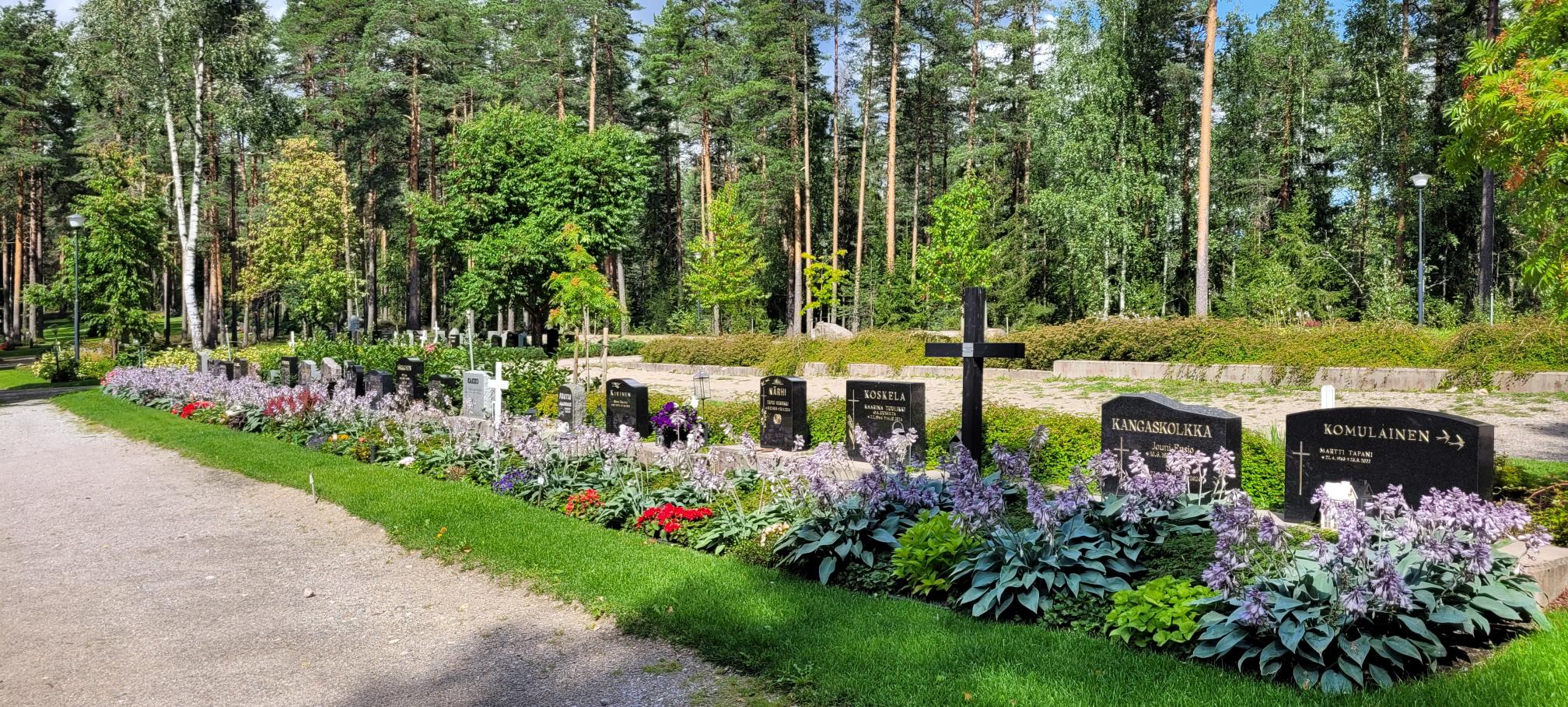 Betonipalkin päällä olevia hautamuistomerkkejä. Hautojen edessä isot perennaistutukset.
