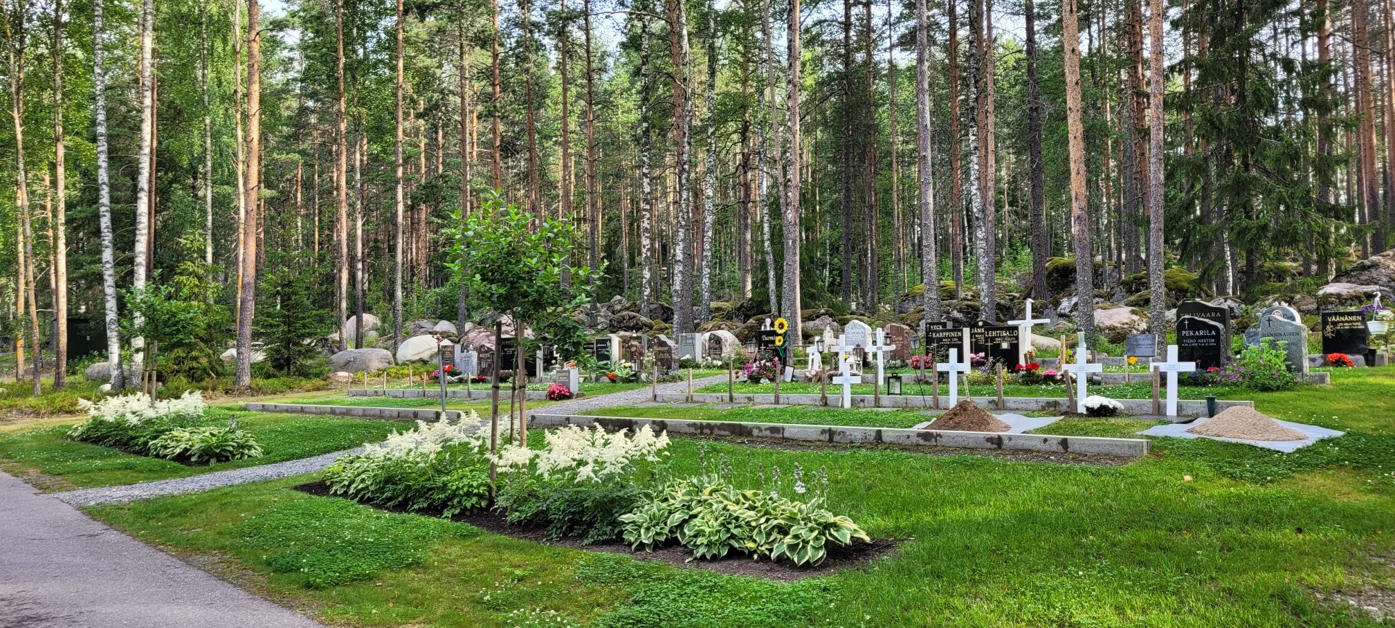 Yhtenäisen aluspalkin päällä muistomerkkejä. Etupuolella perenna- ja pikkupuuistuksia, taustalla metsää.