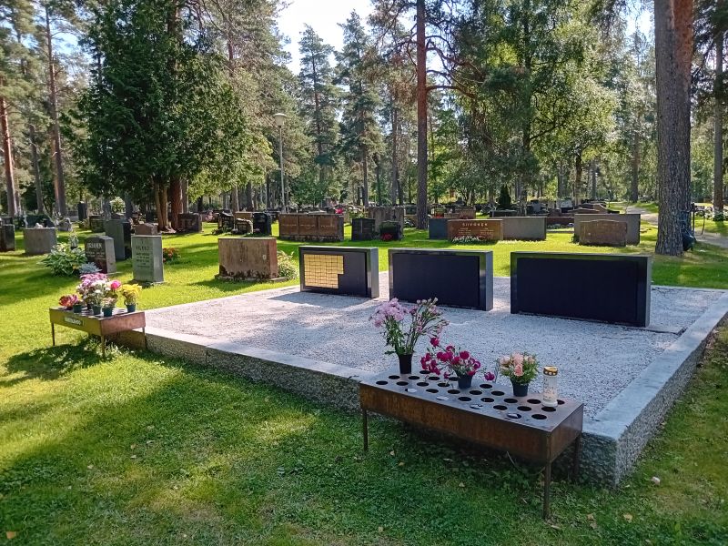 Hiekkapohjainen hauta, kukkapöydät edessä, hautakivet hiekalla.