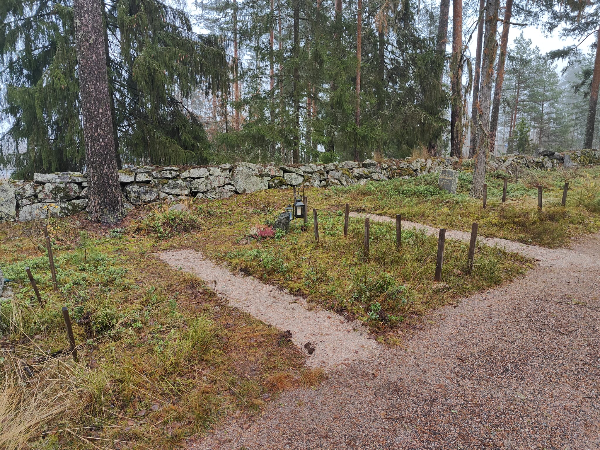 Uurnahautapaikkoja luonnontilaisella metsänpohjalla. Taustalla hautausmaata kiertävää kivimuuria.