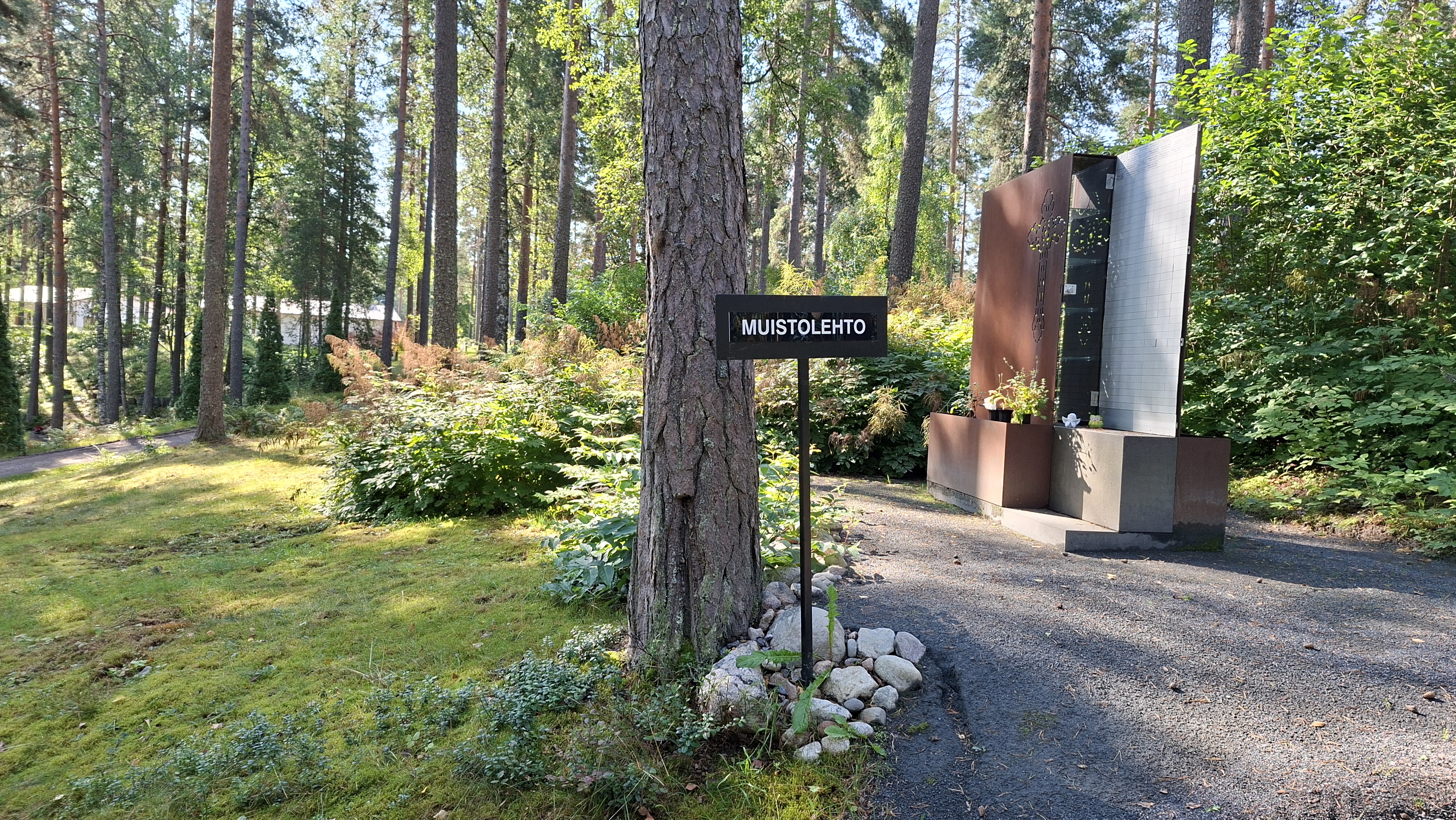 Corten-teräksinen muistomerkki, jossa vainajan nimilaattoja. Ympärillä istutuksia.