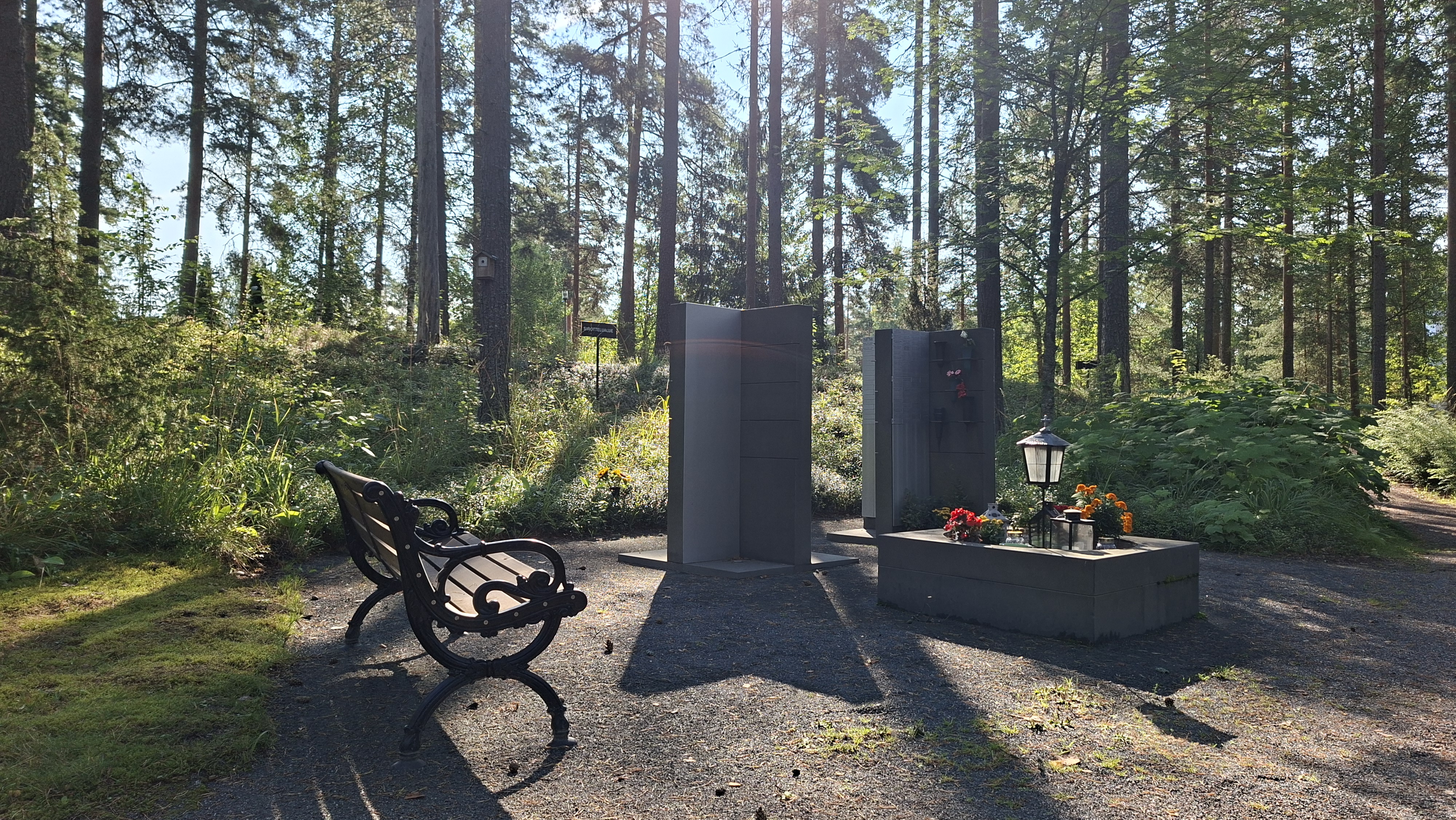 Kaksi korkeaa kivipylvästä vainajan nimilaattoja varten, kukkapöytä, penkki, taustalla mäntymetsää.